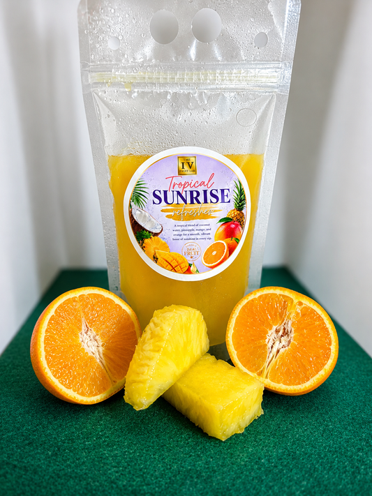 A stand-up pouch of Tropical Sunrise fruit juice filled with bright orange-yellow juice, placed on a dark green surface with sliced orange halves and pineapple chunks in front. The pouch has a clear plastic top and a round tropical fruit label.